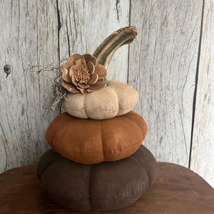May include: A decorative stack of three fabric pumpkins in descending shades of dark brown, medium rust orange, and light beige. The top pumpkin features a natural wooden stem, a dried wooden flower accent, and wispy gray Spanish moss.