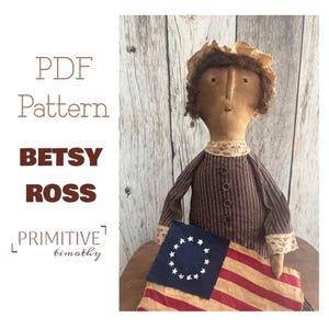 Puede incluir: Un patrón PDF para una muñeca Betsy Ross, una muñeca primitiva que sostiene una bandera con 13 estrellas en un círculo. La muñeca lleva un vestido a rayas con un cuello de encaje y tiene el pelo castaño.