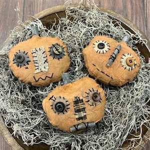 May include: Three handmade, tan fabric pumpkins with stitched details, button eyes, and burlap patches, arranged in a wooden bowl filled with dried moss. The pumpkins have metal stems and are a rustic, fall-themed decoration.
