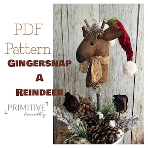 May include: A handcrafted reindeer head with a red Santa hat and a burlap scarf. The reindeer is made of brown fabric and is surrounded by pinecones, dried flowers, and greenery. The text "PDF Pattern GINGERSNAP A REINDEER" is visible.