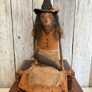May include: A handmade rag doll witch with a black pointed hat, long brown hair, and a brown dress. She is sitting on a wooden box with a black bird perched on her lap.
