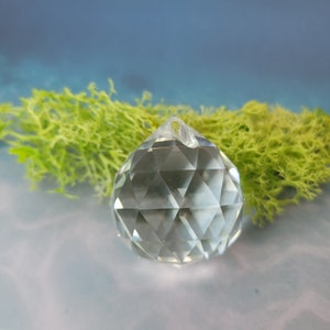 Op de afbeelding: Een heldere, facetgeslepen kristallen bol met een kleine lus aan de bovenkant. Het kristal staat voor felgroen mos en een vage blauw-witte achtergrond. De kristallen bol is een decoratief item.