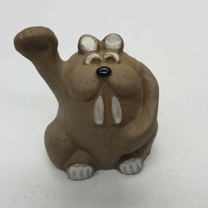 May include: A brown ceramic figurine of a cartoon walrus with white eyes, a black nose, and white tusks. The walrus is waving with its right arm and has white feet.