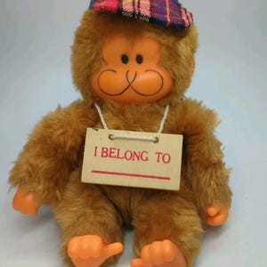 May include: A plush brown monkey toy with orange hands and feet, wearing a plaid hat. The monkey has an orange face with a smiling expression. A wooden tag around its neck reads "I BELONG TO".