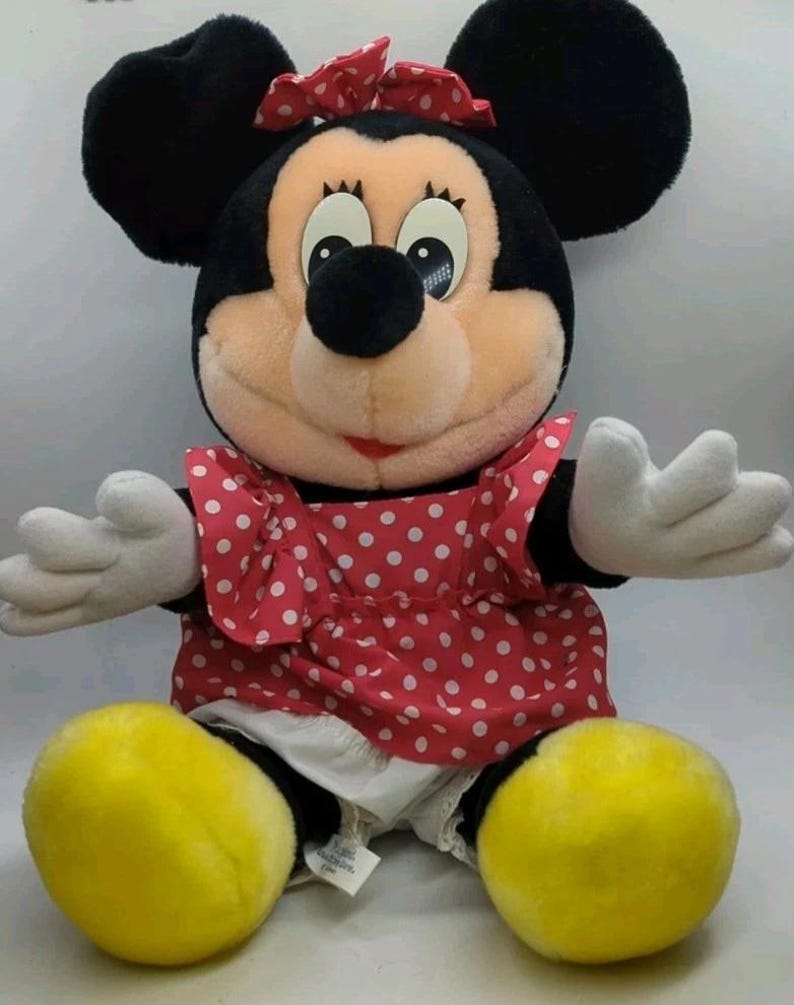 May include: A plush Minnie Mouse doll with black ears, a red polka-dot dress, and a matching bow. The doll has white gloves and yellow shoes. The doll is a classic Disney character.