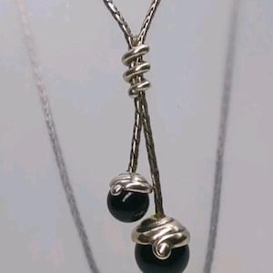 K&ouml;nnte beinhalten: Silberfarbene Halskette mit einzigartigem Design. Die Kette teilt sich in zwei Str&auml;nge, die jeweils mit einer schwarzen, kugelf&ouml;rmigen Perle enden. Die Perlen sind mit silberfarbenem Metall bedeckt. Das zentrale Element ist ein gewickeltes Silbermetall.