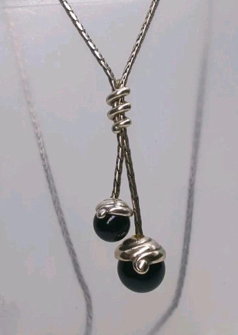 K&ouml;nnte beinhalten: Silberfarbene Halskette mit einzigartigem Design. Die Kette teilt sich in zwei Str&auml;nge, die jeweils mit einer schwarzen, kugelf&ouml;rmigen Perle enden. Die Perlen sind mit silberfarbenem Metall bedeckt. Das zentrale Element ist ein gewickeltes Silbermetall.