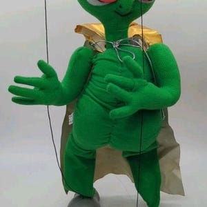 May include: A green alien puppet with a large head, wearing pink-framed sunglasses with blue lenses and a gold cape. The puppet has gray feet and is controlled by strings.