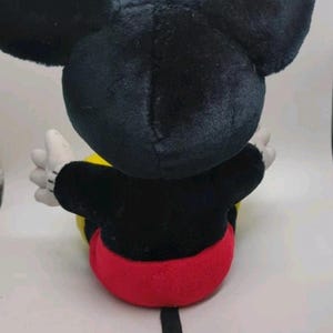 May include: A plush Mickey Mouse toy, viewed from the back. The toy features large black ears, a black body, red shorts, yellow accents, and white gloves. The toy is sitting down.