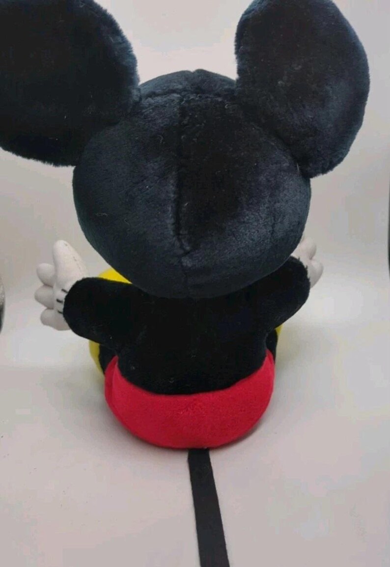 May include: A plush Mickey Mouse toy, viewed from the back. The toy features large black ears, a black body, red shorts, yellow accents, and white gloves. The toy is sitting down.