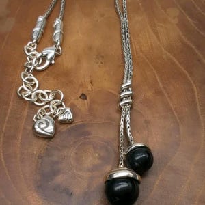 K&ouml;nnte beinhalten: Silberfarbene Halskette mit Y-f&ouml;rmigem Design. Die Halskette hat eine Kette mit zwei Str&auml;ngen, die in schwarzen, runden Perlen mit silbernen Kappen enden. Sie hat eine dekorative Kette mit herzf&ouml;rmigen Anh&auml;ngern und einem Karabinerverschluss.