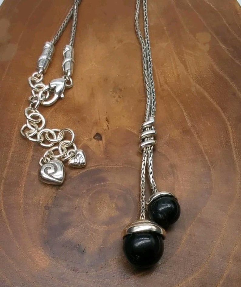 K&ouml;nnte beinhalten: Silberfarbene Halskette mit Y-f&ouml;rmigem Design. Die Halskette hat eine Kette mit zwei Str&auml;ngen, die in schwarzen, runden Perlen mit silbernen Kappen enden. Sie hat eine dekorative Kette mit herzf&ouml;rmigen Anh&auml;ngern und einem Karabinerverschluss.