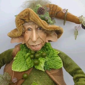 May include: A handcrafted doll featuring a wrinkled face with large ears, wearing a brown velvet hat adorned with a small pinecone, green berries, and dangling leaf accents. The doll wears a green textured shirt with a brown vest and green leaf collar.