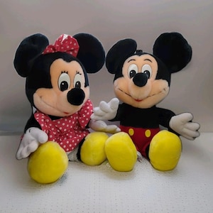 May include: Two plush Disney characters, Mickey and Minnie Mouse, sit side-by-side. Mickey wears red shorts and Minnie a red polka-dot dress and bow. Both have black ears, white gloves, and yellow feet.