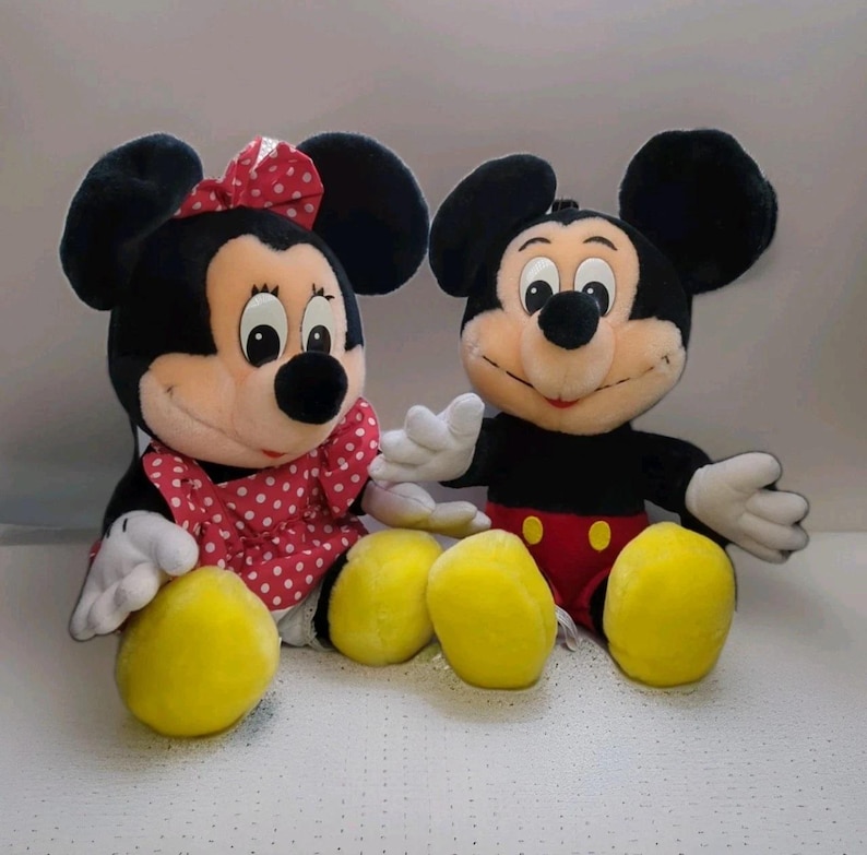 May include: Two plush Disney characters, Mickey and Minnie Mouse, sit side-by-side. Mickey wears red shorts and Minnie a red polka-dot dress and bow. Both have black ears, white gloves, and yellow feet.