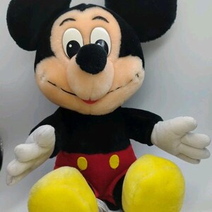 May include: A plush Mickey Mouse toy with black ears, a black body, red shorts with yellow buttons, and white gloves. The toy has a smiling face with a black nose and is sitting with its legs spread.