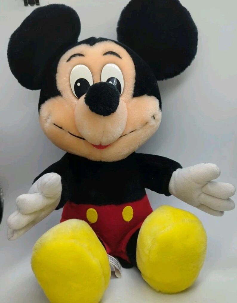 May include: A plush Mickey Mouse toy with black ears, a black body, red shorts with yellow buttons, and white gloves. The toy has a smiling face with a black nose and is sitting with its legs spread.