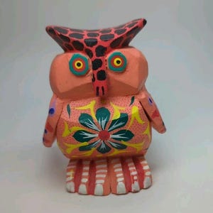 May include: A hand-carved wooden owl figurine. The owl is painted in shades of coral, red, black, yellow, and green. The owl's chest features a floral design. The owl's eyes are yellow with blue and green accents. The owl's feet are painted with white and red stripes.
