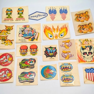 May include: A collection of vintage stickers with various designs. The stickers feature illustrations of tigers, dragons, skulls, and other cartoonish characters. Some stickers have text like "Goodyear", "Spider", and "Look Ma, No Cavities!"