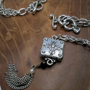 Peut inclure: Gros plan sur des bijoux argent&eacute;s sur une surface en bois brun. Les bijoux comprennent un bracelet &agrave; cha&icirc;ne avec un fermoir &agrave; bascule, une cha&icirc;ne avec un charme en forme de c&oelig;ur et un pendentif carr&eacute; avec des d&eacute;tails orn&eacute;s. Une gland argent&eacute; et une perle noire sont &eacute;galement visibles.