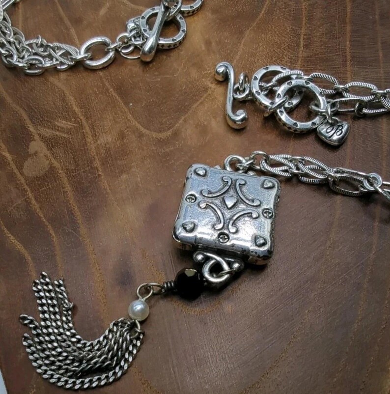 Peut inclure: Gros plan sur des bijoux argent&eacute;s sur une surface en bois brun. Les bijoux comprennent un bracelet &agrave; cha&icirc;ne avec un fermoir &agrave; bascule, une cha&icirc;ne avec un charme en forme de c&oelig;ur et un pendentif carr&eacute; avec des d&eacute;tails orn&eacute;s. Une gland argent&eacute; et une perle noire sont &eacute;galement visibles.