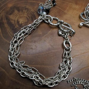 Peut inclure: Bracelet argent&eacute; avec plusieurs rang&eacute;es de cha&icirc;nes et un fermoir &agrave; bascule. Le bracelet comporte un charm d&eacute;coratif avec des perles bleues et noires et une perle. Le bijou est pr&eacute;sent&eacute; sur une surface en bois.
