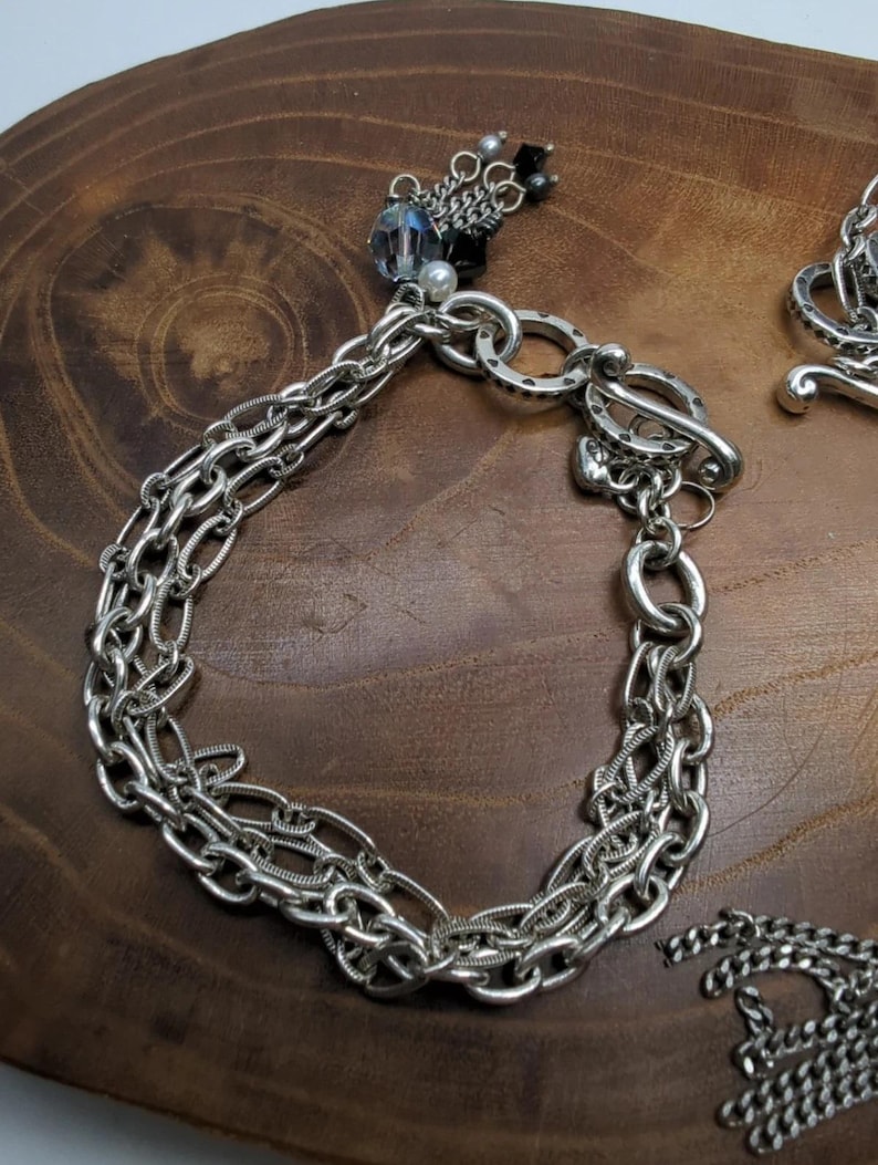Peut inclure: Bracelet argent&eacute; avec plusieurs rang&eacute;es de cha&icirc;nes et un fermoir &agrave; bascule. Le bracelet comporte un charm d&eacute;coratif avec des perles bleues et noires et une perle. Le bijou est pr&eacute;sent&eacute; sur une surface en bois.