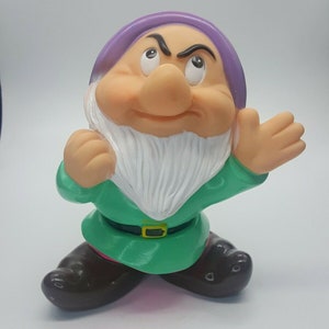 May include: A purple-hatted, green-clad, grumpy-looking dwarf figurine with a white beard. The dwarf is standing with his arms outstretched, one hand raised in a gesture of annoyance.