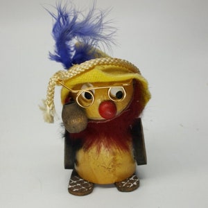 May include: A wooden figurine of a person wearing a yellow hat with a blue feather, round glasses, and a red nose. The figurine is holding a pipe in its mouth and has a red and brown fur collar.