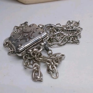 Peut inclure: Un pendentif argent&eacute; de forme rectangulaire avec des volutes d&eacute;coratives sur le couvercle, attach&eacute; &agrave; une cha&icirc;ne. La cha&icirc;ne est constitu&eacute;e de maillons entrelac&eacute;s. Le pendentif est ferm&eacute;, peut-&ecirc;tre un m&eacute;daillon.