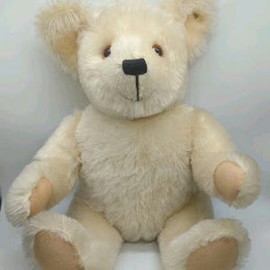 May include: A cream-colored plush teddy bear with soft, shaggy fur, featuring a black stitched nose and mouth, brown eyes, and smooth, light tan fabric pads on the soles of its feet and palms of its hands, presented in a seated position.