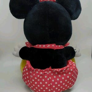 May include: A plush Minnie Mouse toy, viewed from the back, featuring large black ears and a red polka-dot skirt. The toy has white gloves and a red polka-dot bow. The overall design is a classic representation of the iconic character.