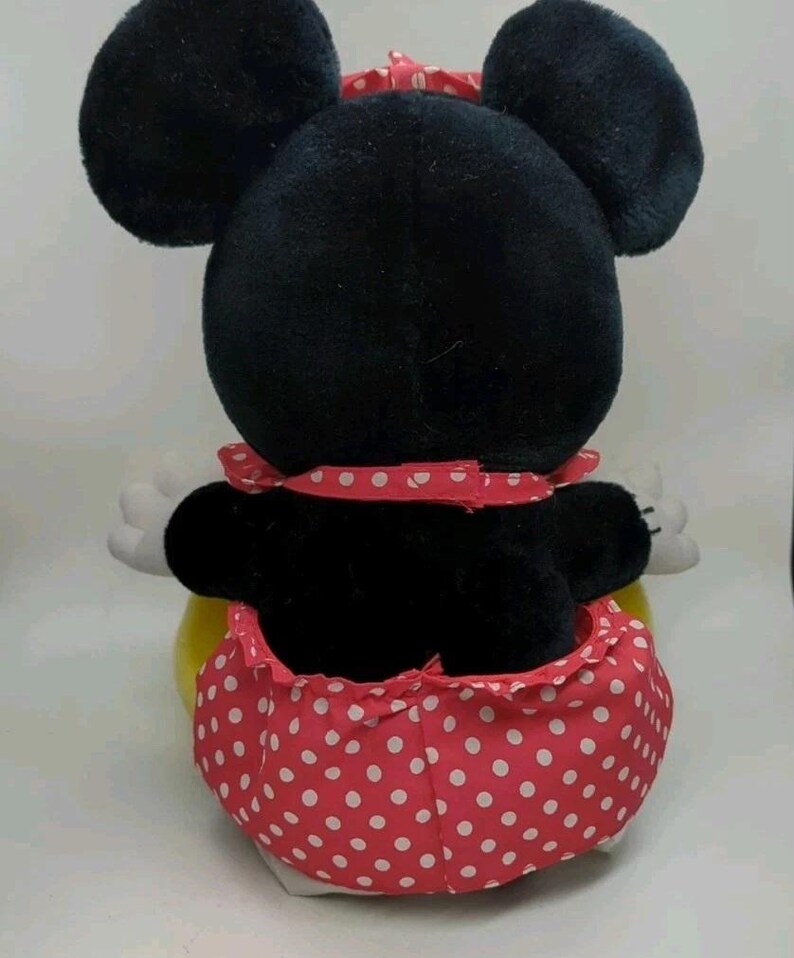 May include: A plush Minnie Mouse toy, viewed from the back, featuring large black ears and a red polka-dot skirt. The toy has white gloves and a red polka-dot bow. The overall design is a classic representation of the iconic character.