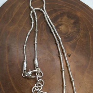 K&ouml;nnte beinhalten: Eine silberne Halskette mit detailliertem Kettendesign. Die Halskette verf&uuml;gt &uuml;ber einen Karabinerverschluss und eine Reihe dekorativer Glieder. Die Kette besteht aus kleinen, miteinander verbundenen Segmenten, die ein strukturiertes Aussehen erzeugen. Die Halskette wird auf einer Holzoberfl&auml;che pr&auml;sentiert.