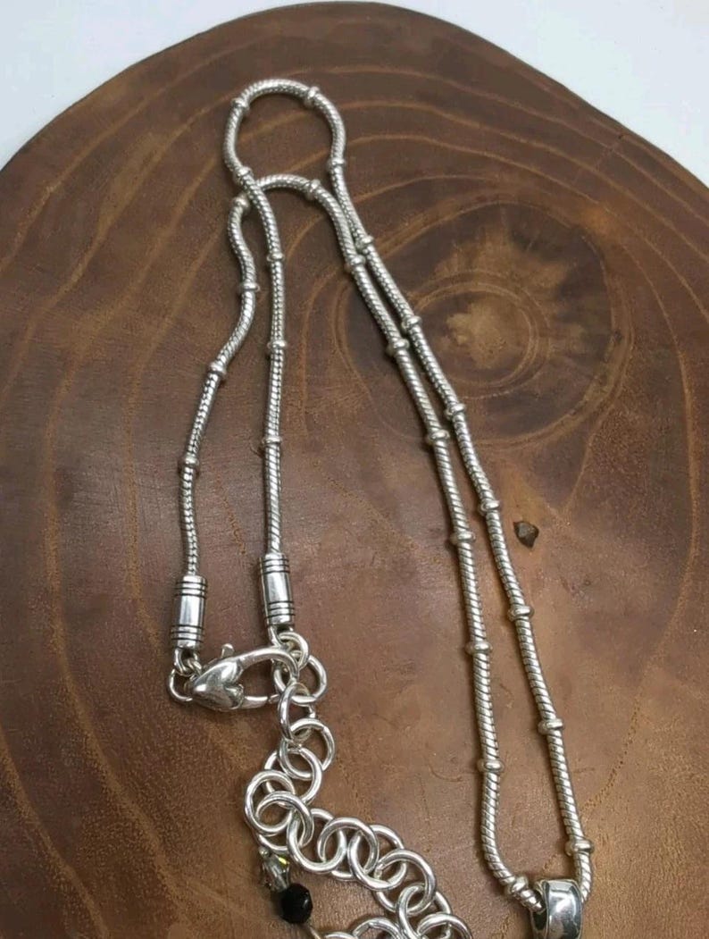 K&ouml;nnte beinhalten: Eine silberne Halskette mit detailliertem Kettendesign. Die Halskette verf&uuml;gt &uuml;ber einen Karabinerverschluss und eine Reihe dekorativer Glieder. Die Kette besteht aus kleinen, miteinander verbundenen Segmenten, die ein strukturiertes Aussehen erzeugen. Die Halskette wird auf einer Holzoberfl&auml;che pr&auml;sentiert.