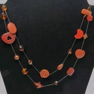 May include: A two-strand necklace featuring orange and amber-colored beads. The necklace includes various shapes, such as hearts, teardrops, and geometric forms. The beads are strung on a thin, silver-colored wire, creating a layered effect.