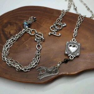 Peut inclure: Un ensemble de bijoux argent&eacute;s comprenant un bracelet et un collier avec un pendentif en forme de c&oelig;ur. Le bracelet a un fermoir &agrave; bascule, et le collier a un pendentif carr&eacute; d&eacute;coratif avec un c&oelig;ur d&eacute;coup&eacute;. L'ensemble comprend un pompon et de petites perles.