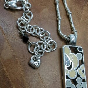 K&ouml;nnte beinhalten: Silberfarbene Halskette mit einem rechteckigen Anh&auml;nger mit einem floralen Design in Schwarz, Creme und Grau. Die Halskette enth&auml;lt einen herzf&ouml;rmigen Anh&auml;nger mit Initialen und eine Kette mit herzf&ouml;rmigem Verschluss.