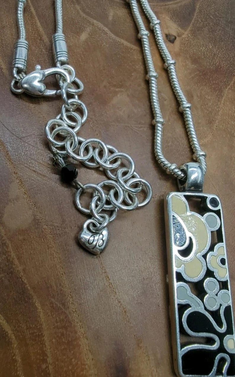 K&ouml;nnte beinhalten: Silberfarbene Halskette mit einem rechteckigen Anh&auml;nger mit einem floralen Design in Schwarz, Creme und Grau. Die Halskette enth&auml;lt einen herzf&ouml;rmigen Anh&auml;nger mit Initialen und eine Kette mit herzf&ouml;rmigem Verschluss.