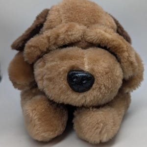 May include: A plush toy dog in shades of brown. The dog has floppy ears, a black plastic nose, and a soft, furry texture. The toy is designed to resemble a puppy and is likely intended for children.