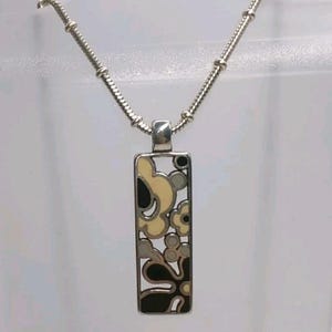 K&ouml;nnte beinhalten: Silberfarbene Halskette mit Anh&auml;nger, rechteckiger Anh&auml;nger mit floralem Design in Schwarz, Creme und Grau. Die Halskette hat eine filigrane Kette mit Perlenstruktur. Der Anh&auml;nger ist ca. 3,8 cm lang.