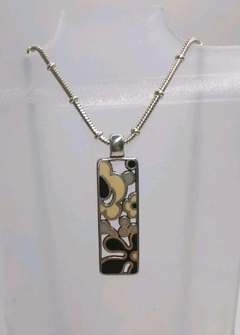 K&ouml;nnte beinhalten: Silberfarbene Halskette mit Anh&auml;nger, rechteckiger Anh&auml;nger mit floralem Design in Schwarz, Creme und Grau. Die Halskette hat eine filigrane Kette mit Perlenstruktur. Der Anh&auml;nger ist ca. 3,8 cm lang.