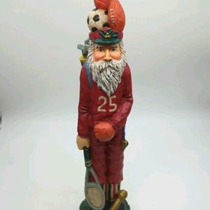 May include: A red Santa Claus figurine wearing a sports-themed hat with a soccer ball, football, and other sports equipment. The figurine has a white beard, a red jersey with the number 25, and holds a tennis racket and a dumbbell. The base is green.