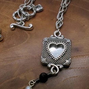 Peut inclure: Collier argent&eacute; avec un pendentif carr&eacute; orn&eacute; d'un c&oelig;ur &eacute;vid&eacute;. Le pendentif a une bordure textur&eacute;e et est reli&eacute; &agrave; une cha&icirc;ne. Un fermoir &agrave; bascule est visible. Une pampille de cha&icirc;nes et de perles argent&eacute;es pend en dessous.