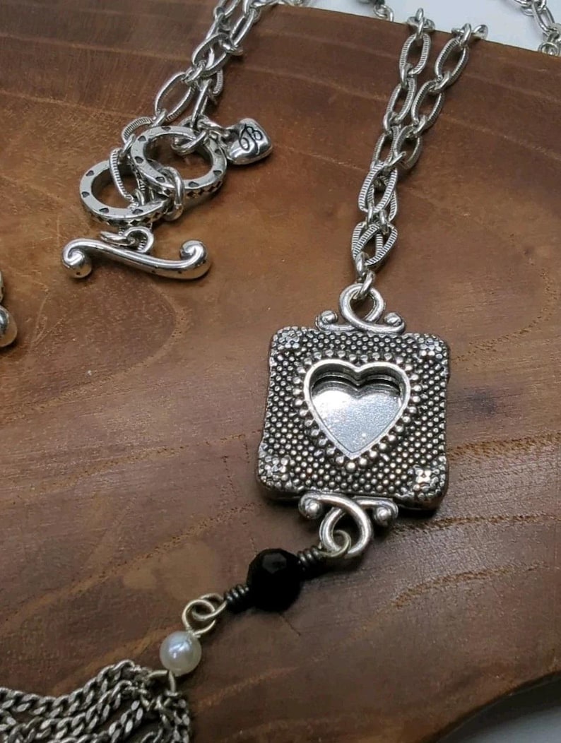 Peut inclure: Collier argent&eacute; avec un pendentif carr&eacute; orn&eacute; d'un c&oelig;ur &eacute;vid&eacute;. Le pendentif a une bordure textur&eacute;e et est reli&eacute; &agrave; une cha&icirc;ne. Un fermoir &agrave; bascule est visible. Une pampille de cha&icirc;nes et de perles argent&eacute;es pend en dessous.