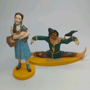 May include: A figurine of a girl with brown hair in braids, wearing a blue and white checkered dress, holding a small brown basket and a gray dog. Beside her is a scarecrow figurine in a green coat, sitting on a yellow base with a black crow.