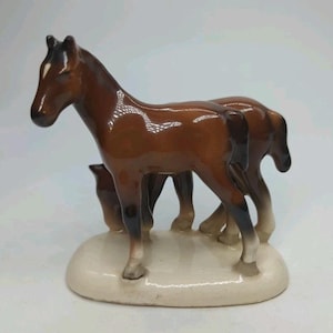 May include: A ceramic figurine of two brown horses standing side-by-side on a cream-colored base. The horses have dark brown manes and tails, with a white marking on the forehead of the lead horse. The figurine is glossy.