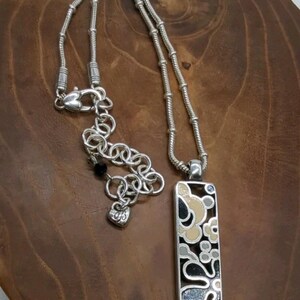 K&ouml;nnte beinhalten: Silberfarbene Halskette mit rechteckigem Anh&auml;nger mit floralem Design in Schwarz, Beige und Grau. Die Halskette hat eine Kette mit Karabinerverschluss und eine verstellbare Kette. Der Anh&auml;nger ist ca. 3,8 cm lang.