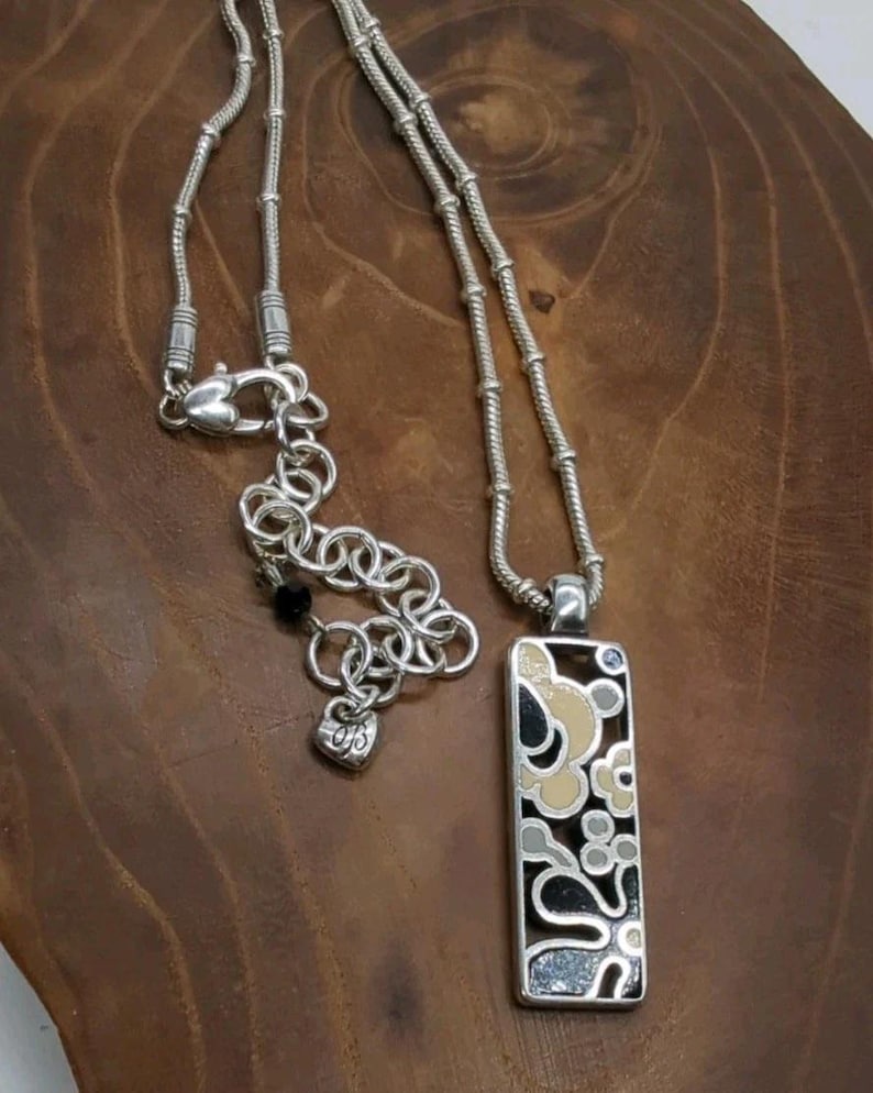 K&ouml;nnte beinhalten: Silberfarbene Halskette mit rechteckigem Anh&auml;nger mit floralem Design in Schwarz, Beige und Grau. Die Halskette hat eine Kette mit Karabinerverschluss und eine verstellbare Kette. Der Anh&auml;nger ist ca. 3,8 cm lang.