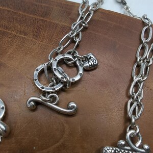 Peut inclure: Gros plan sur un collier argent&eacute; avec un fermoir &agrave; bascule. Le collier pr&eacute;sente divers maillons de cha&icirc;ne et &eacute;l&eacute;ments d&eacute;coratifs, dont un petit charme au design orn&eacute;. Le fond est une surface en bois.